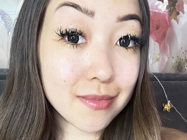 RenyLime has pale skin, brown eyes, long brown hair, and is smiling with glossy pink lips. She has thick black eyelashes and a small beauty mark on her right cheek.