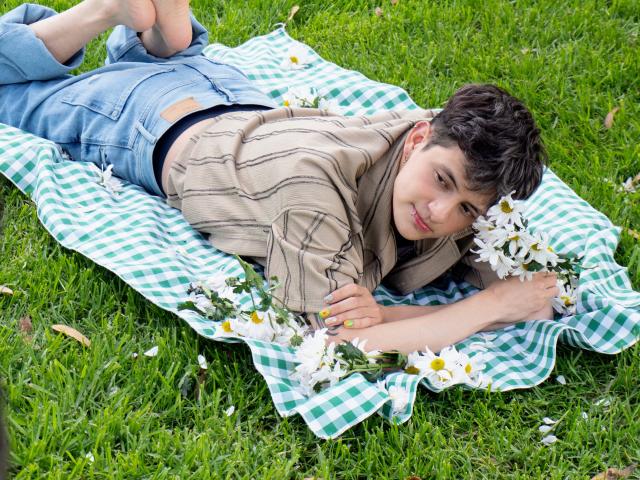 GabyThoms lies on a green checkered blanket, wearing a beige striped shirt and blue jeans, holding daisies, with bare feet up, on grass, smiling at the camera.