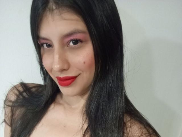 MadissonFontaine has long black hair, pink eyeshadow, red lipstick, and a small beauty mark on her left cheek. She's smiling with her boobs barely visible in the top.