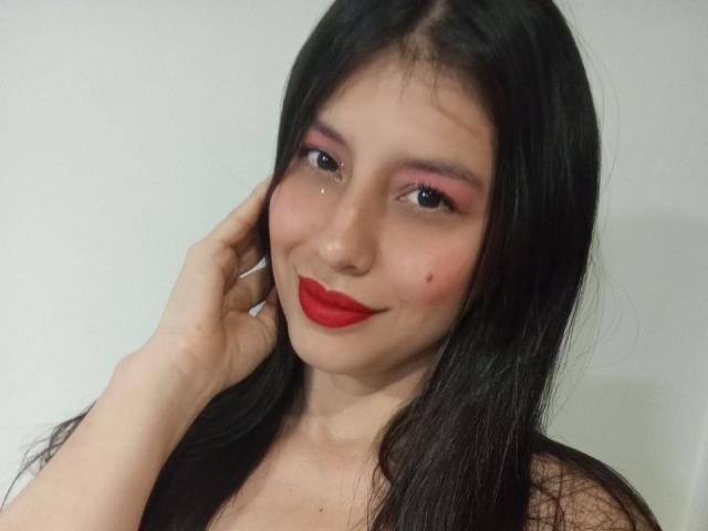 MadissonFontaine has long black hair, pink eye makeup, red lipstick, and a cute smile with a small beauty mark on her left cheek. She's touching her hair.