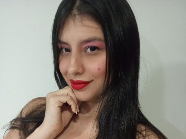 MadissonFontaine has long black hair, red lipstick, pink eyeshadow, and a small mole on her cheek, smiling with her hand near her face, showing her nude shoulders.