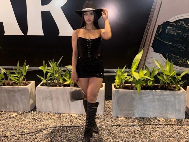 LaurentRay, gekleed in een zwarte mini-jurk zonder bandjes met knopen, zwarte laarzen en een cowboyhoed, met een grijze handtas, staat voor groene planten op grind..