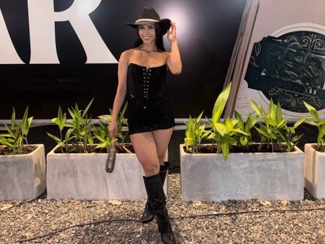 LaurentRay con un trasero grande, un corsé negro, una falda corta, botas negras, un sombrero de vaquero, sosteniendo un cinturón, plantas verdes, grava, sonriendo. Está de pie, con una mano en el sombrero, en un entorno bien iluminado..