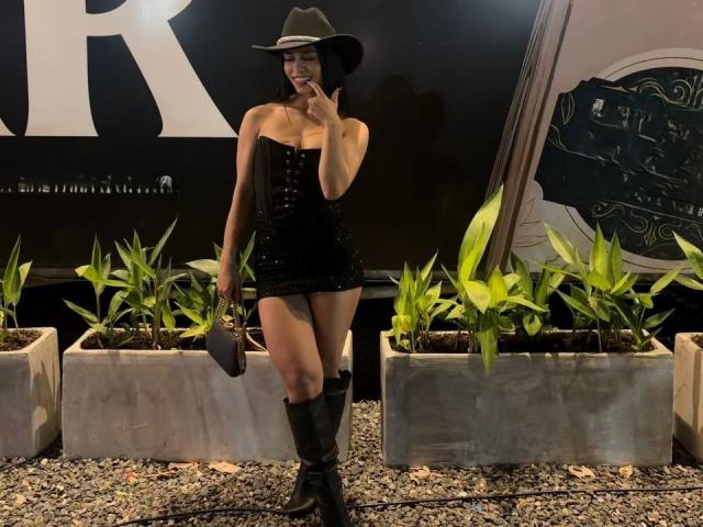 LaurentRay, vêtue d'un corset noir sans bretelles, d'une mini-jupe, d'un chapeau de cowboy noir, de bottes montantes, tenant une pochette grise, debout avec un doigt sur les lèvres, plantes vertes au premier plan..