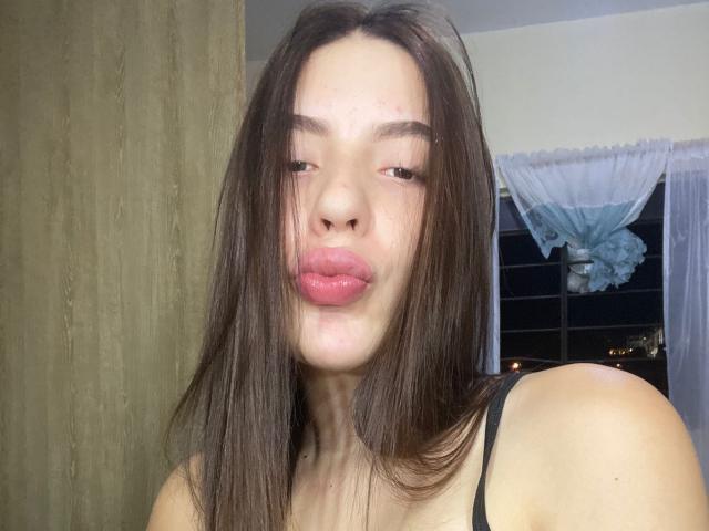 Mannuela69, con cabello castaño largo, labios rosados y gruesos, ojos ligeramente entrecerrados y una camiseta negra, está de pie frente a una pared de madera, luciendo adorable..