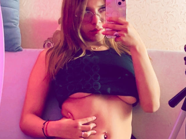 HeraJuno with glasses, lifting a black top to expose her small tits and belly button piercing, holding a phone in a mirror selfie. Her hand rests on her flat stomach.