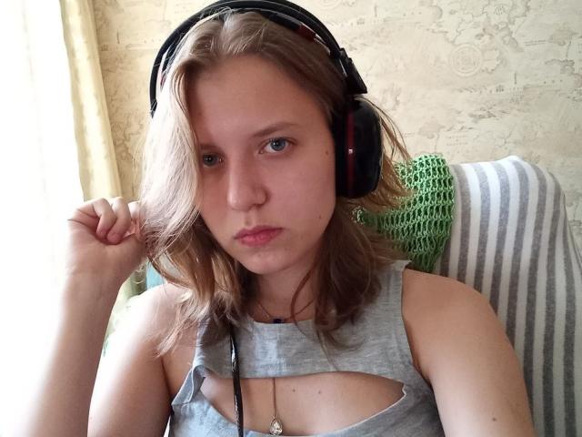 VikiCharm con una camiseta gris, con un corte que muestra pequeños pechos, usando auriculares, cabello castaño claro, expresión de labios fruncidos, mano cerca de la oreja, almohada de ganchillo verde, silla a rayas, piel pálida..