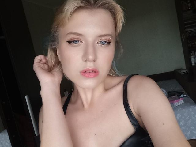 VikiCharm, avec ses cheveux blonds, ses yeux bleus et ses lèvres roses, porte un soutien-gorge noir, une main dans les cheveux, et regarde l'appareil photo. Ses seins sont visibles, mais les mamelons ne sont pas montrés..