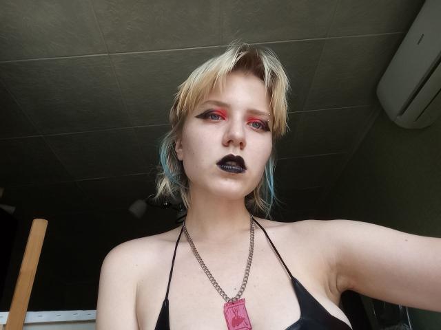 VikiCharm com cabelo loiro curto, com mechas vermelhas e azuis, batom preto, delineador vermelho, top de alça cavada preto, colar com pingente vermelho, pele clara e axila visível, olhando para a câmera..