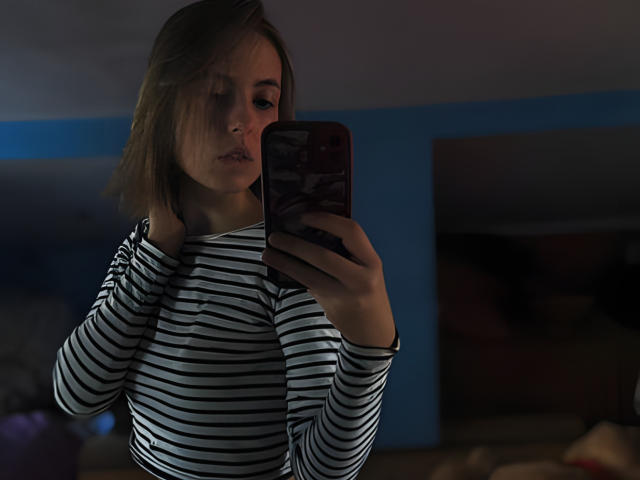 Tora Stambaugh, avec des cheveux bruns mi-longs, portant une chemise à manches longues rayée, tenant un téléphone avec une coque rouge, prenant un selfie devant un miroir, les yeux partiellement fermés, regardant vers le bas..