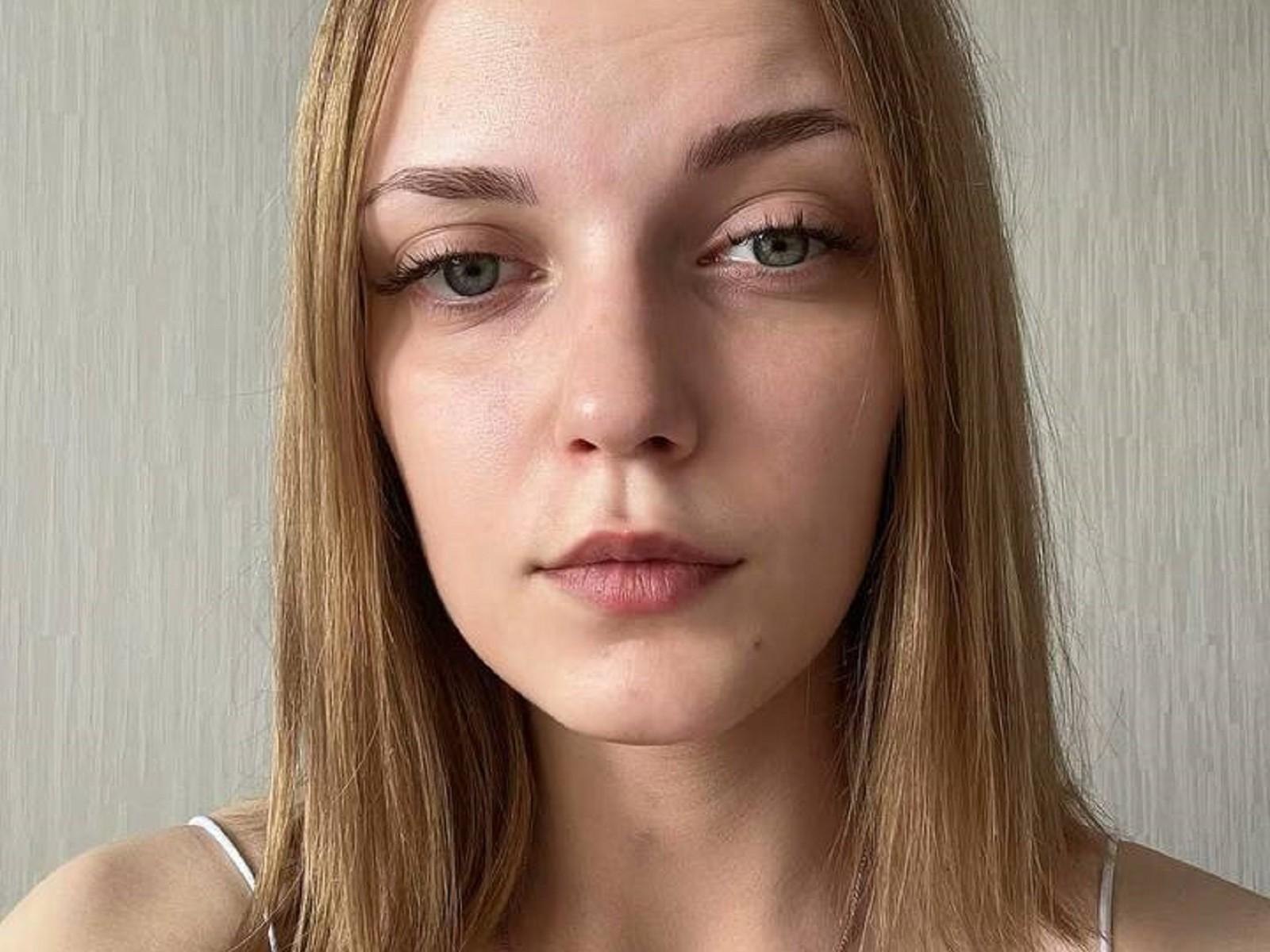LeighaSetters a les cheveux blonds et lisses, des yeux verts et une expression neutre. Elle porte un haut blanc à bretelles fines, et son nez est légèrement retroussé. Ses lèvres sont légèrement rosées..