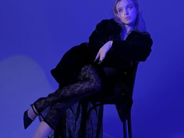 RosendaLazcano with blue lighting, wearing black lace skirt, black high heels, black fur coat, and a serious expression, sitting on a chair with one leg crossed.