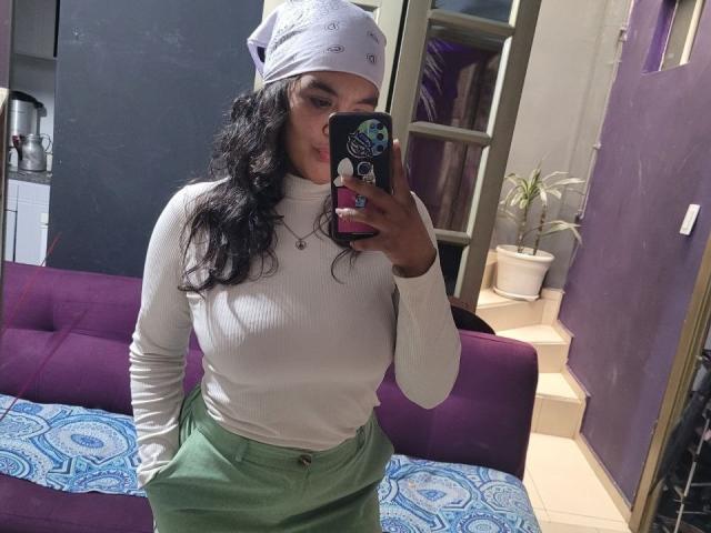 ZiaNoir with curly black hair, white beanie, white ribbed turtleneck, green pants, holding phone in mirror selfie, showing her tits and ass. Room has purple sofa and potted plant.