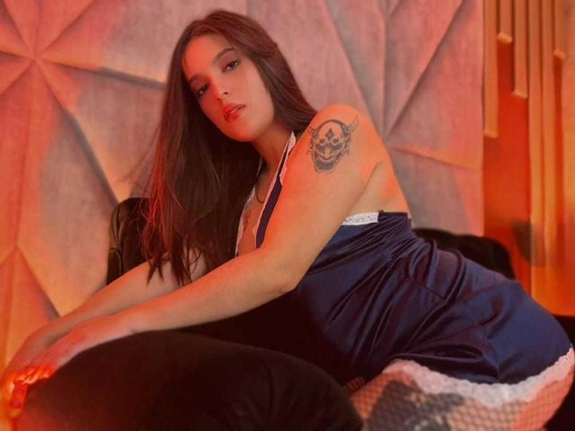 Katarina Novak mit langem, braunen Haar, rotem Lippenstift, blauem Seidenkleid, weißer Spitzenbordüre, Tattoo am linken Arm, sitzend auf einem schwarzen Kissen, blickt in die Kamera, warmes, oranges Licht..