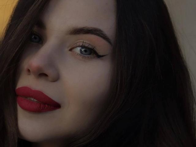 O rosto de AzzieStelman, com cabelo castanho escuro, olhos azuis, delineador preto e batom vermelho, está ligeiramente virado para o lado. Sua pele é clara e ela está usando maquiagem escura..