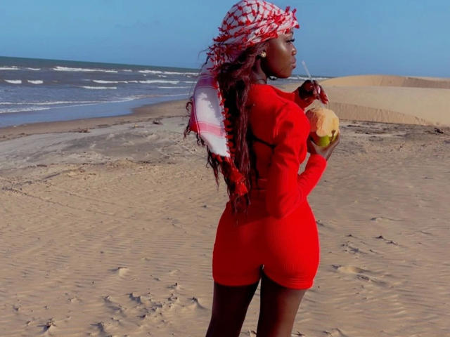 EbonyAngel-hot in red dress, holding coconut, with red and white checkered scarf, standing on sandy beach, looking back, long red braids, dark skin, blue sky, ocean in background.