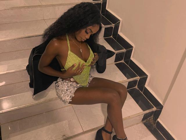 EbonyAngel-morena, com pele escura e cabelos pretos cacheados, vestindo uma blusa verde de renda e shorts prateados, sentada nos degraus, tocando seus pequenos seios brilhantes, com uma jaqueta preta pendurada sobre ela..