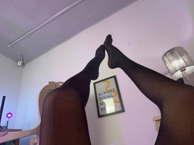 IrisBlair's black pantyhose-covered legs are raised, showing her toes pointing at the ceiling. She's wearing sheer black stockings, with a white wall and lamp in the background.