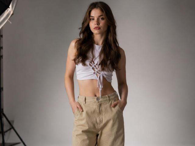 KristianSeidel a de longs cheveux bruns, un débardeur blanc avec un laçage sur le devant, et un pantalon beige avec les mains dans les poches, debout avec une expression neutre, regardant la caméra..