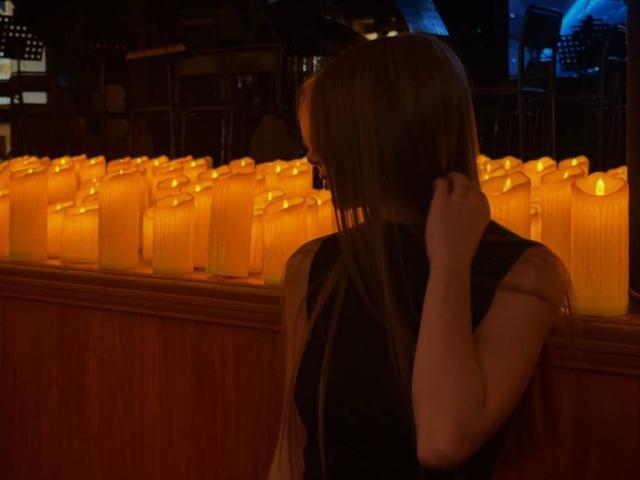 LesterKassner with long brown hair, black dress, and hand in hair, standing by orange-lit candles, facing away, in dimly lit room. Boobs partially visible.
