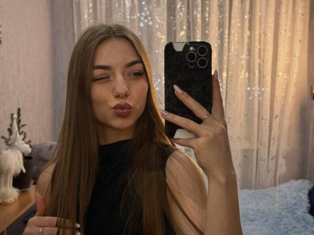 LesterKassner with long brown hair, pouty lips, wearing a black top, holding a phone, taking a mirror selfie, showing off her nails and ring, in a cozy room.