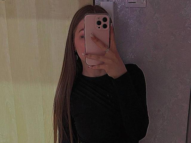 LesterKassner with long brown hair, black long-sleeve shirt, holding pink phone covering face, reflection in mirror, wearing ring on right hand, pale skin, in dimly lit room.