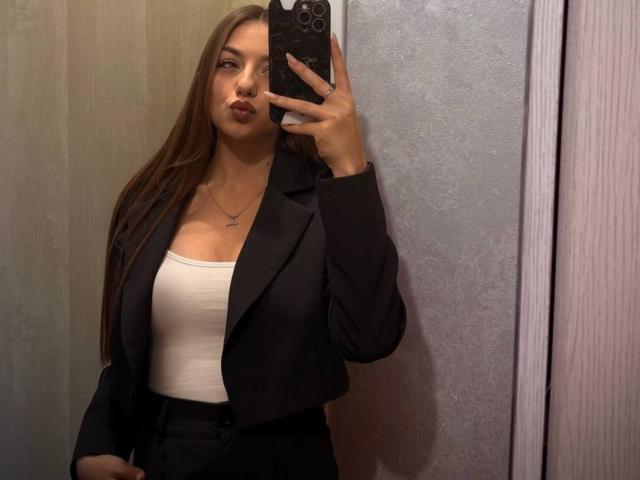 LesterKassner with long brown hair, pouty lips, wearing a black blazer over a white tank top, holding a phone, showing her tits, standing in a mirror selfie.