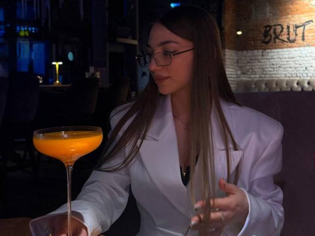 LesterKassner, wearing a white coat, glasses, and long brown hair, holds a glass of orange cocktail in one hand and a wine glass in the other.