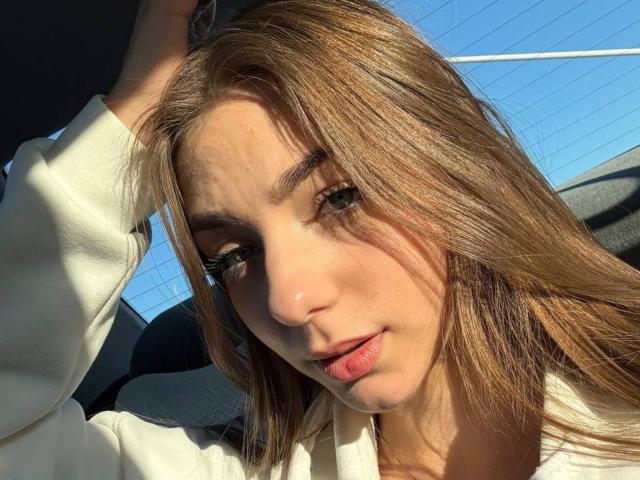 LesterKassner with long brown hair, light skin, and pouty pink lips, wearing a white hoodie, hand in hair, sunlight highlighting face, blue sky in background.