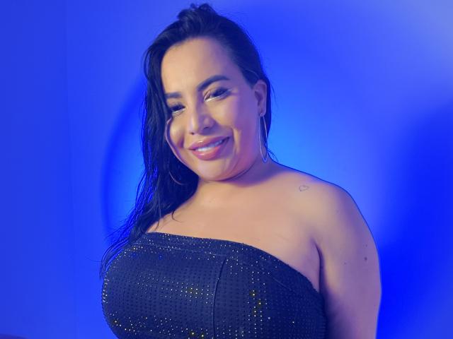 Nahomi Rosse mit nassen, schwarzen Haaren, lächelnd, trägt ein schwarzes, trägerloses Kleid mit Pailletten, Reifenohrringe und ein kleines Herz-Tattoo an ihrer rechten Schulter, vor einem blauen Hintergrund..