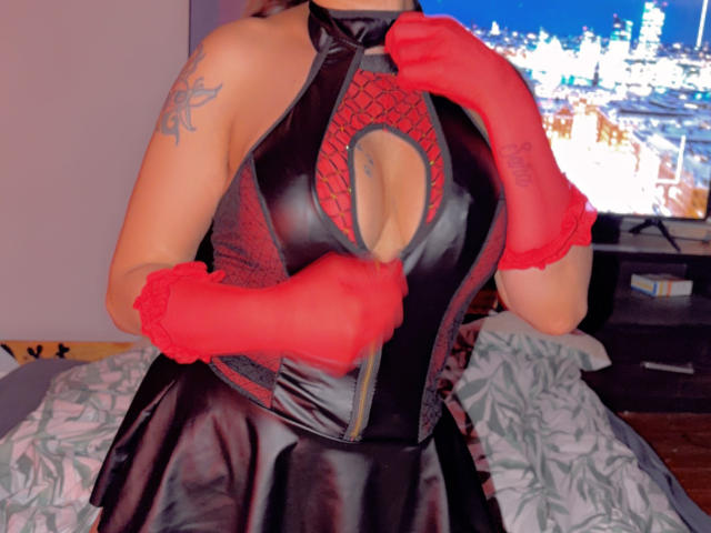 Saraby portant une robe en latex noire avec une découpe, des gants en dentelle rouge, montrant ses seins, un tatouage sur l'épaule gauche, tenant la robe, debout dans une chambre avec vue sur la ville..