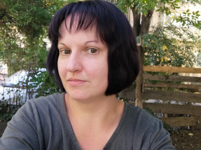 LadyAlma, avec ses cheveux noirs courts, ses yeux marrons et son t-shirt gris à col en V, a une expression neutre, avec un léger froncement de sourcils, et est assise à l'extérieur..