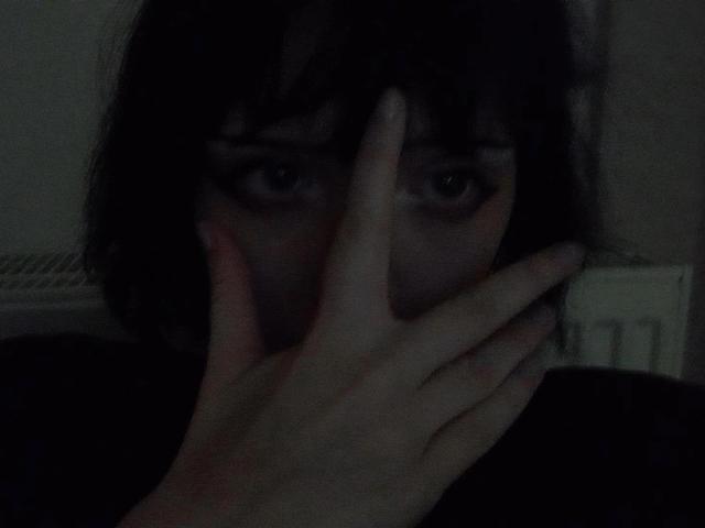 NikaDreamOne, com cabelos escuros e desalinhados, cobrindo a boca com a mão, mostrando seus olhos em uma fotografia escura e com pouca luz. Seus dedos parcialmente escondem seu nariz e boca..