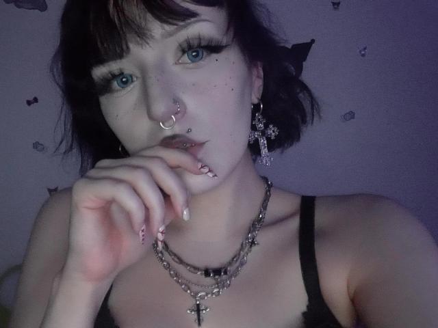 NikaDreamOne has pale skin, blue eyes, black hair, and multiple piercings, including a nose ring and lip piercing. She's wearing black lingerie with a chain necklace.