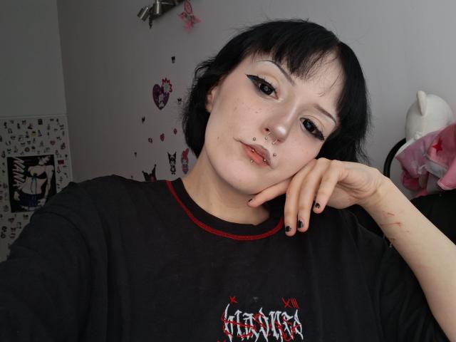 NikaDreamOne, com cabelo preto, maquiagem gótica nos olhos, piercing no nariz, piercing nos lábios, camisa preta e unhas pretas, com a cabeça apoiada na mão, olhando para a câmera. Ela é fofa, com um leve semblante de descontentamento..