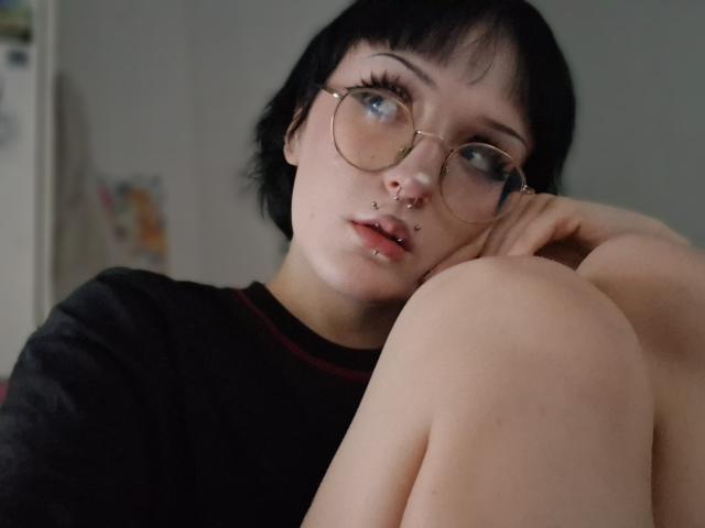 NikaDreamOne met kort zwart haar, een bril, een neusring, een lip-piercing en een zwart shirt, zittend met de knieën opgetrokken, kijkend naar de camera met een licht geopende mond..