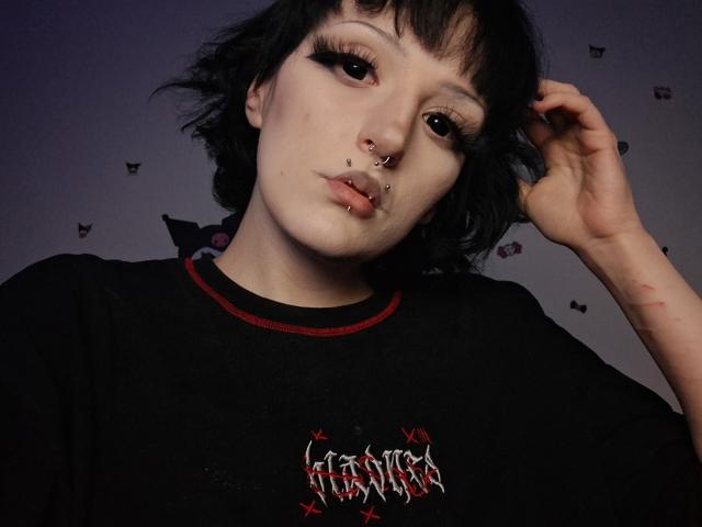 NikaDreamOne with black short hair, nose ring, lip ring, and dark makeup, wearing a black shirt with red trim, raising her hand, showing a slight smirk.