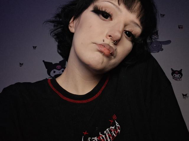 NikaDreamOne heeft kort zwart haar, donkere eyeliner, een neusring, een septumring en een lipring. Ze draagt een zwart shirt met rode accenten en kijkt naar beneden met een lichte frons..