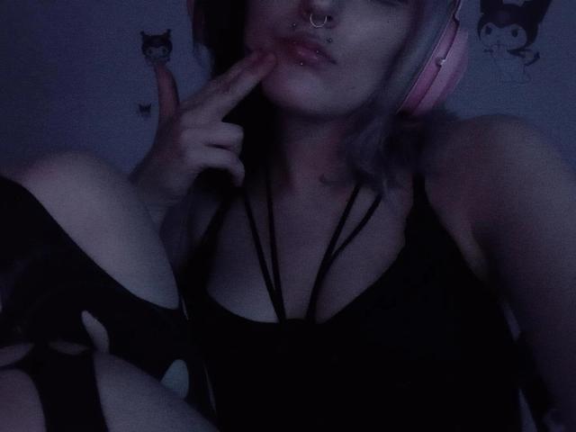 NikaDreamOne met roze hoofdtelefoon, zwart strapless topje, vinger in de mond, neusring, bleke huid, roze haar, zittend, benen gekruist, foto met donkerblauwe tint. Borsten, decolleté, licht zichtbaar..