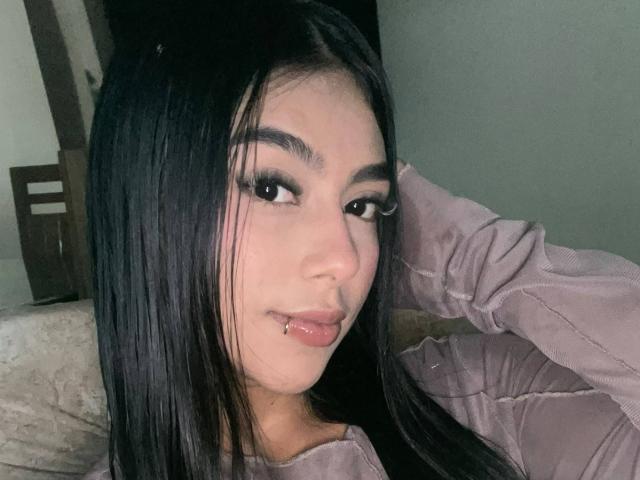 SophiaBrownn, met lang zwart haar, draagt een roze gewatteerde jas, heeft een lip-piercing en kijkt met een lichte grijns naar de camera. Haar ogen zijn zwaar omlijnd..