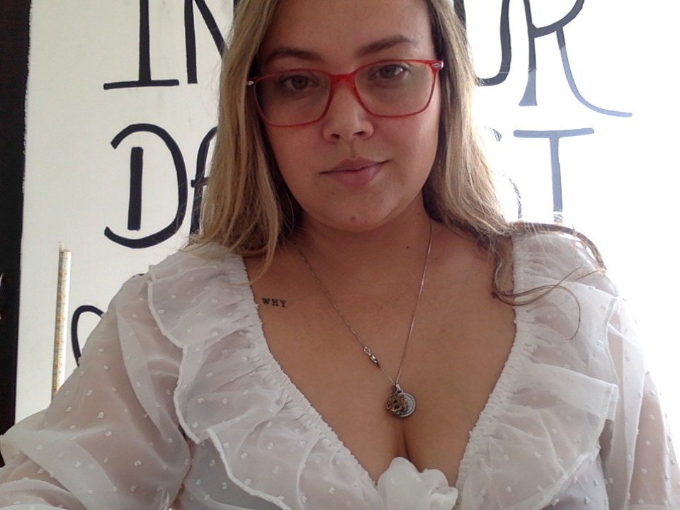 Brennda69, com óculos vermelhos, blusa branca com bolinhas, decote profundo, colar de prata pequeno, cabelo loiro, pele clara, sorriso discreto, tatuagem no lado direito do peito. Seios visíveis sob a blusa..