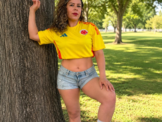 EmiliaMilf está de pie frente a un árbol, vestida con una camiseta de fútbol amarilla con detalles en rojo y azul, y pantalones cortos vaqueros, mostrando su abdomen y una sonrisa segura y sensual..