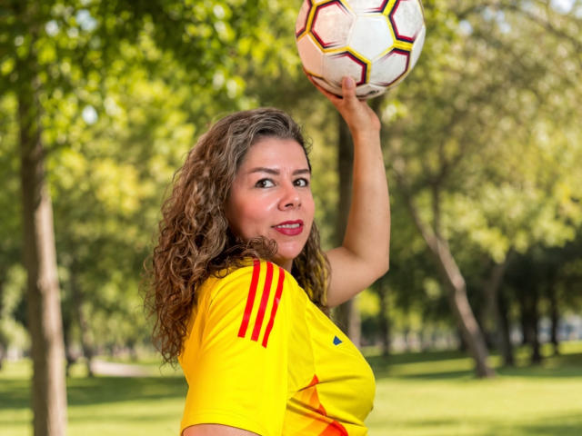 EmiliaMilf, com cabelos castanhos cacheados, camisa de futebol amarela com listras vermelhas, segurando uma bola de futebol acima da cabeça, sorrindo com batom vermelho em um parque ensolarado..