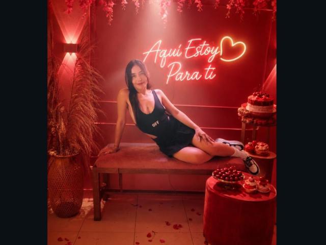 MindyLove with dark hair, medium tits, in a black dress, lying on a red bench, neon heart sign, potted plant, and red table with chocolate-covered strawberries.