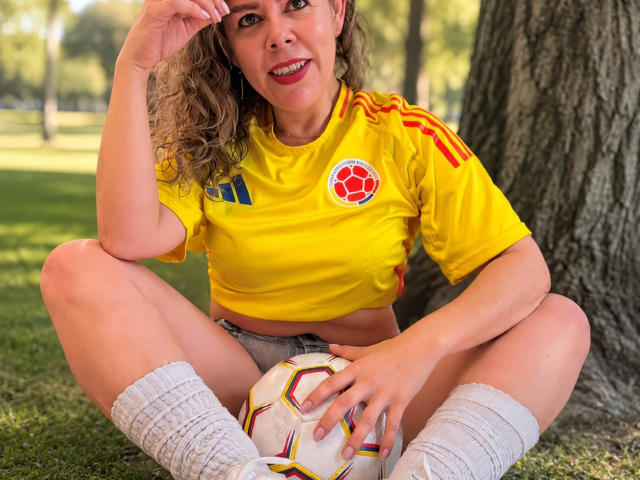 EmiliaMilf, seduta sull'erba, indossa una maglietta da calcio gialla con strisce rosse e gialle, tiene una palla da calcio, calze bianche, capelli biondi ricci, sorride, si tocca la testa, pantaloncini, un albero sullo sfondo..