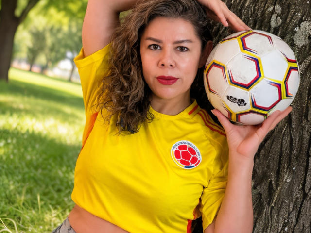 EmiliaMilf with curly brown hair, yellow soccer shirt, red soccer ball, holding it against a tree, looking at camera, red lips, green grass, tree trunk in background.