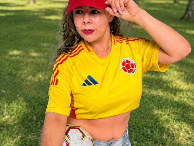 EmiliaMilf wearing a red cap, yellow sports shirt with red and blue details, exposing her midriff, and holding a soccer ball, sitting on grass in a park.