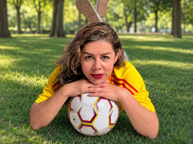 EmiliaMilf está deitada na grama com os seios em cima de uma bola de futebol, vestindo uma camisa de futebol amarela e olhando para a câmera com batom vermelho..