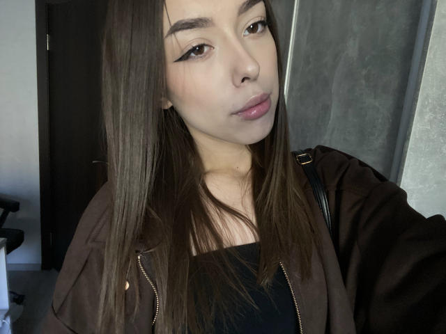 Leila, avec ses longs cheveux bruns, ses yeux bruns et ses lèvres roses, portant un haut noir et une veste marron, se montre dans un selfie, mettant en valeur sa petite poitrine et son joli visage..