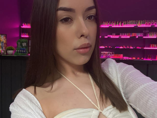 LeilaAllie, met lang bruin haar, lichte huid en volle lippen, draagt een witte top, waarbij haar borsten zichtbaar zijn. Ze kijkt naar de camera in een roze verlichte bar..
